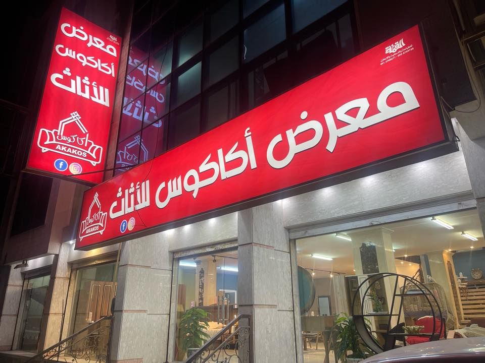 واجهة معرض أكاكوس للأثاث ليلًا، بلافتتها الحمراء وشعار أكاكوس، وقطع أثاث خلف الزجاج